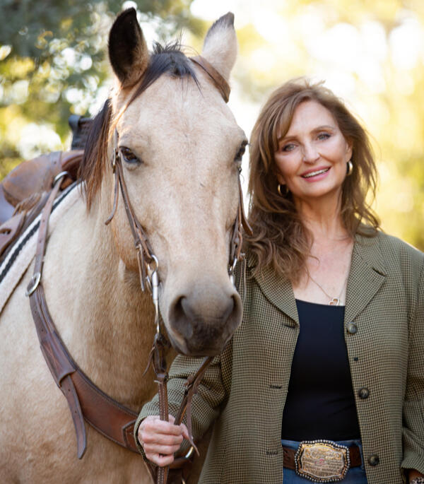 Photo of a woman holding a horse's bridle. The woman is smiling and dressed in business casual dress, but is wearing a large, Western belt buckle.